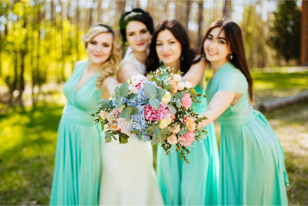 Four bridesmaids in matching turquoise dresses smiling and holding a vibrant floral bouquet outdoors.