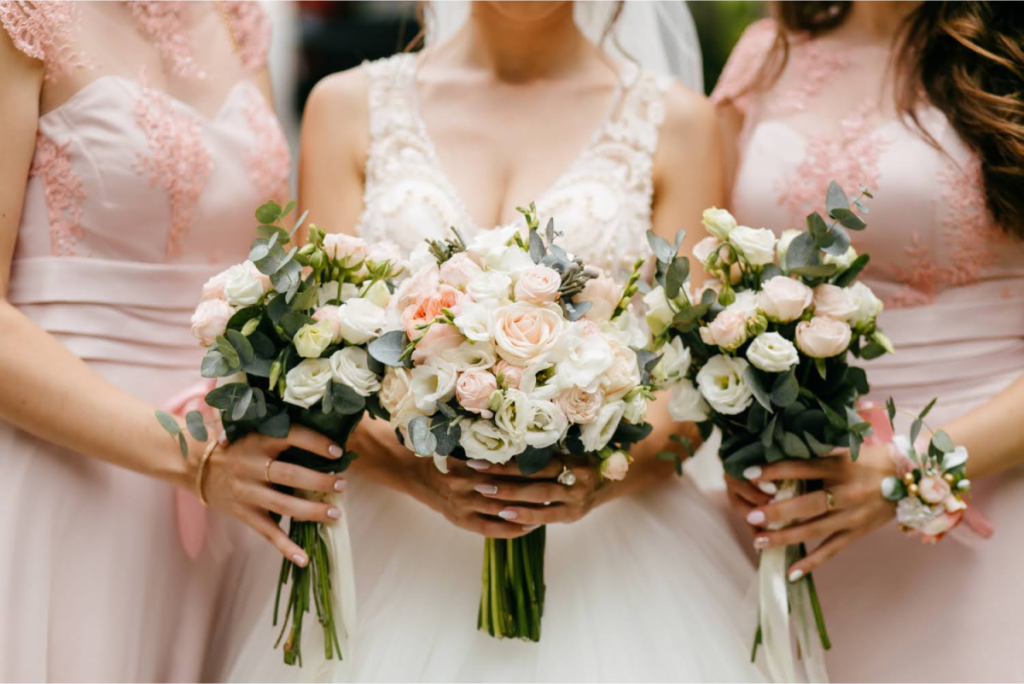 Bride holding a bouquet of white and pink roses, standing between two bridesmaids in matching pink dresses, each holding similar floral arrangements.