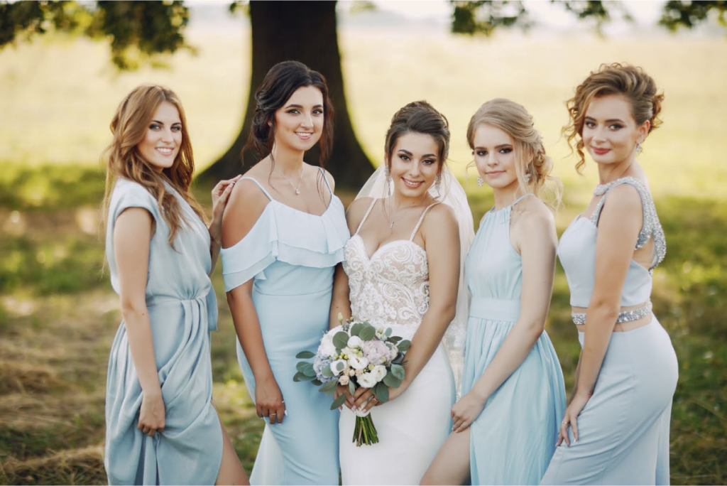 Bride in a white gown standing outdoors with four bridesmaids in light blue dresses, all smiling and posing together.